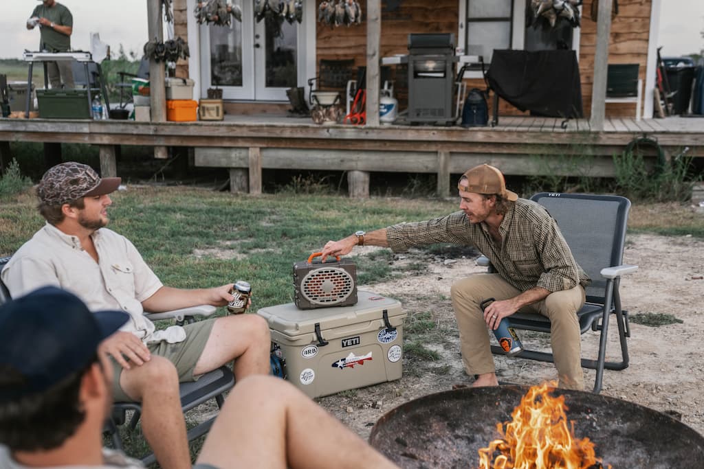 A lifestyle photo of two men sitting by a fire pit with a cooler, surrounded by outdoor equipment. #speaker-color_delta
