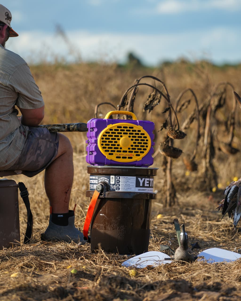 A lifestyle photo of a person sitting in a field with a YETI cooler and purple and yellow device. #speaker-color_purple-gold-gameday-series