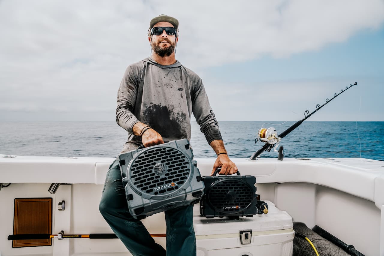 A lifestyle photo of a man in a fishing boat holding a Turtlebox Grande and a Turtlebox Original speakers in gray color #speaker-color_gray