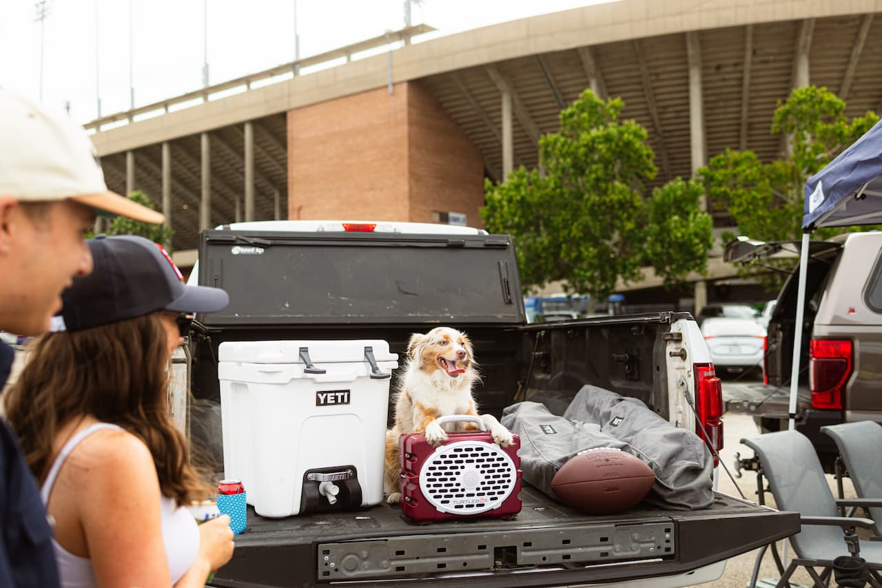 A lifestyle photo of people with a dog in the back of a truck with a Turtlebox Original in Maroon & White and sports equipment. #speaker-color_maroon-white-gameday-series