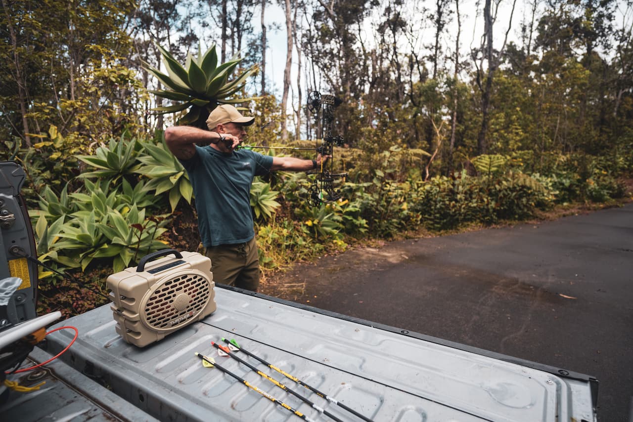 A lifestyle photo of a man shooting a bow  with a tan Turtlebox on the back of his truck  #speaker-color_tan